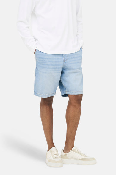 Lichtblauwe jeansshort van ONLY & SONS® met steekzakken, gecombineerd met een wit shirt met lange mouwen en crèmekleurige sneakers.
