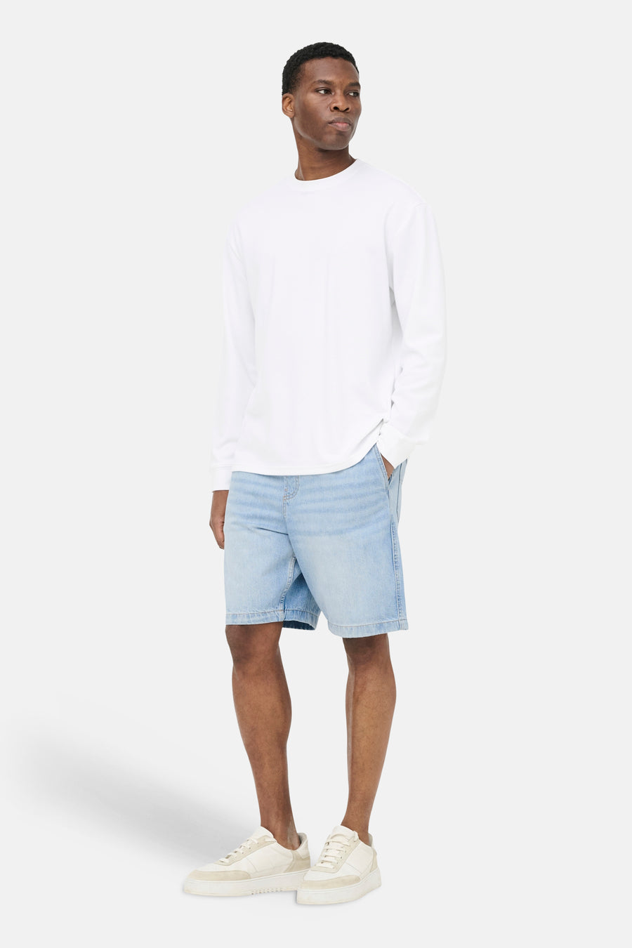 Lichtblauwe denim jeansshort van ONLY & SONS®, gecombineerd met een wit shirt met lange mouwen en crèmekleurige sneakers.
