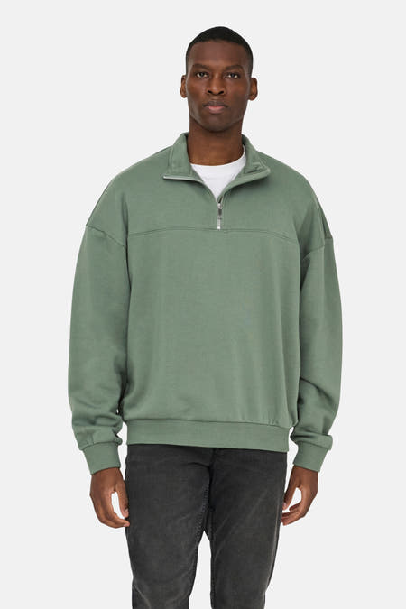 Pull-over vert de ONLY & SONS avec une fermeture à glissière courte et une coupe décontractée, porté avec un shirt blanc et des jeans foncés.