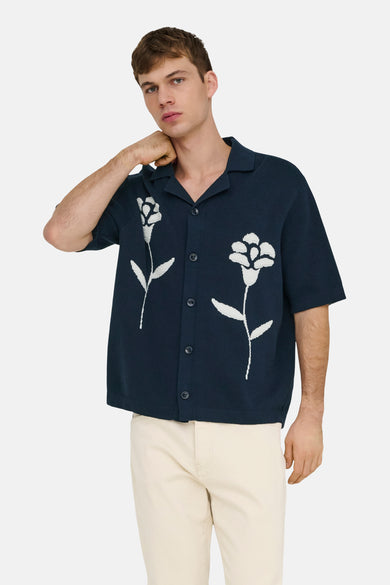 Pull bleu ONLY & SONS® à manches courtes avec motif floral blanc, porté avec un pantalon de couleur crème et des baskets.