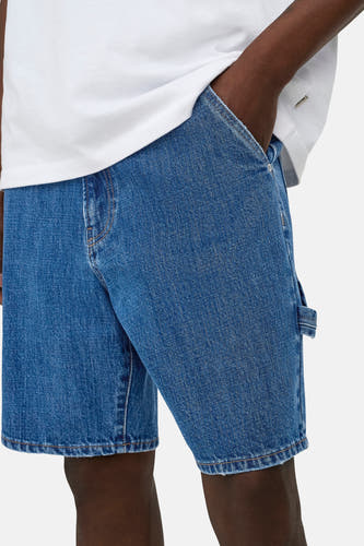 Short en jean bleu moyen ONLY & SONS® avec passant de marteau, porté avec un t-shirt blanc et une main dans la poche.