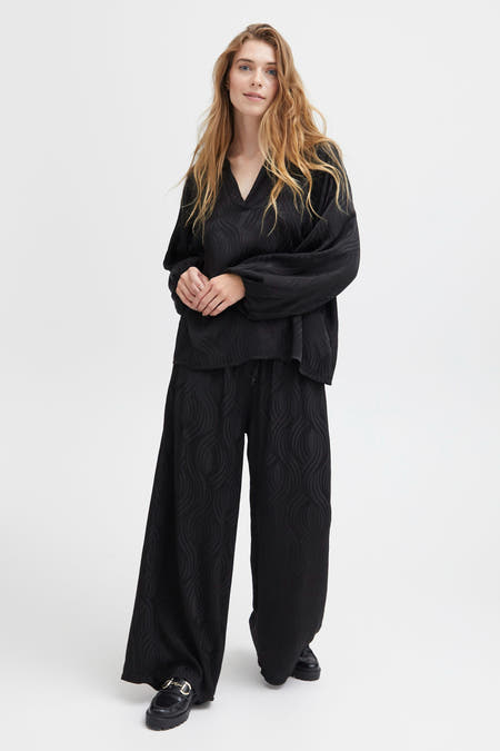 Blouse noire de SORBET avec des manches longues et un motif subtil, associée à une jupe large et ajustée et des chaussures noires.