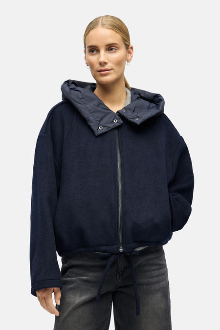 Blauwe korte jas van OBJECT, met capuchon en een trekkoord aan de onderkant, gedragen over een grijze top en een zwarte broek.
