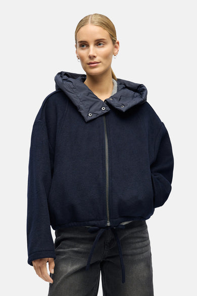 Manteau court bleu d'OBJECT, avec capuche et cordon de serrage à la base, porté sur une chemise grise et un pantalon noir.
