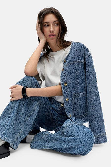 Vrouw zit gekruist in blauwe tweed broek en jas, witte top, zwarte armband en schoenen.