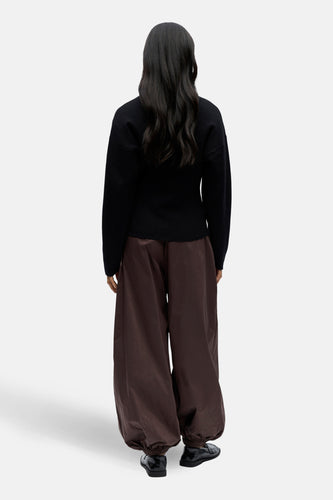 Achterkant van een vrouw in een zwarte trui en wijde, bruine broek, met zwarte schoenen op een witte achtergrond.
