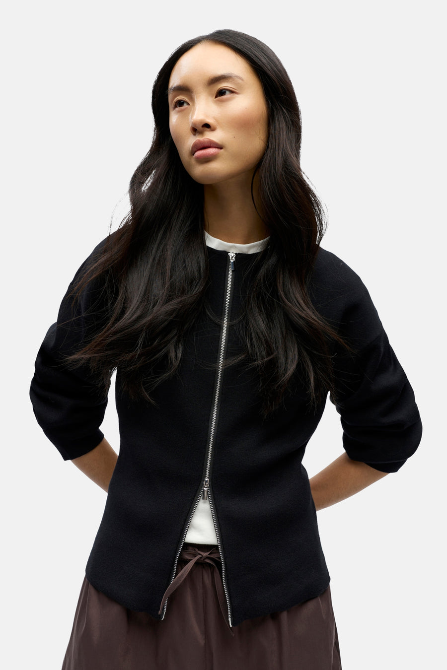 Vrouw met zwarte jas met rits, bruine broek, staand.