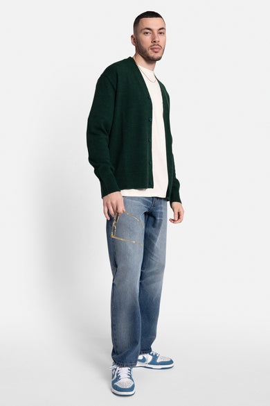 Veste verte de REDEFINED REBEL portée sur un T-shirt crème, avec des jeans bleus et des baskets.