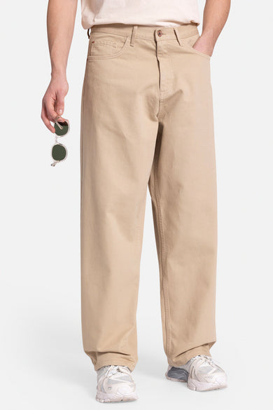 Beige broek van REDEFINED REBEL, losse pasvorm, met zonnebril in de hand en witte sneakers.
