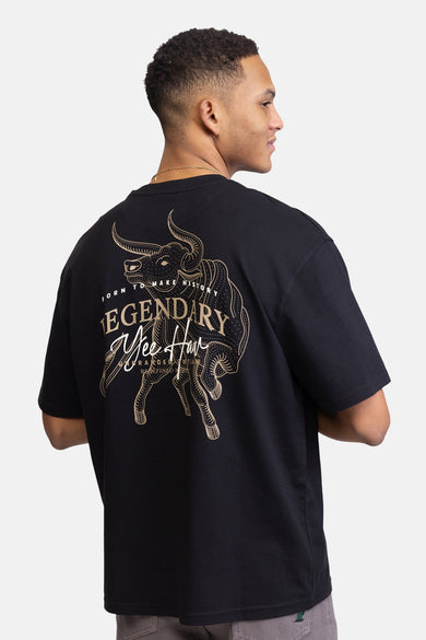 Man met zwarte T-shirt met stierenprint, grijze broek, gouden ketting, staande.