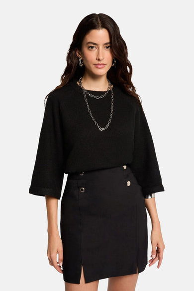 Jupe courte noire de Morgan De Toi, avec des boutons décoratifs et des fentes à la base, associée à une veste noire et des bijoux argentés.