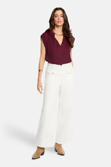 Chemisier bordeaux sans manches de Morgan De Toi, avec un col en V et combiné avec un pantalon blanc et des bottines marron.