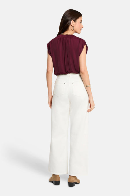 Mouwloze bordeauxrode blouse van Morgan De Toi met plooien, gecombineerd met witte broek en bruine enkellaarsjes, achteraanzicht.
