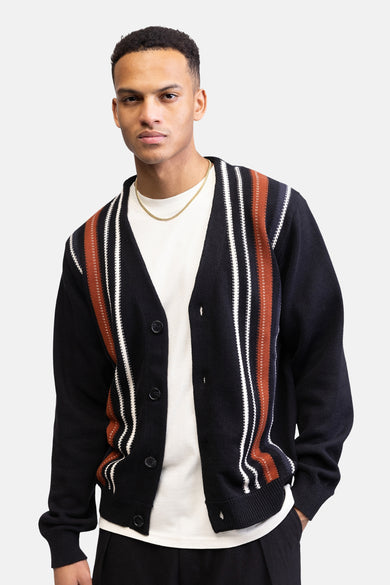 Gilet noir de REDEFINED REBEL, avec des rayures verticales en blanc et marron, porté sur un t-shirt blanc avec une chaîne dorée.