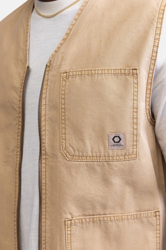 Manteau sans manches beige de REDEFINED REBEL avec poche brodée et fermeture éclair, associé à un t-shirt blanc et une chaîne dorée.