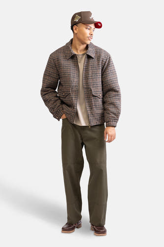 Groene broek van REDEFINED REBEL, gecombineerd met een bruine jas met ruitpatroon, een T-shirt en bruine schoenen.

