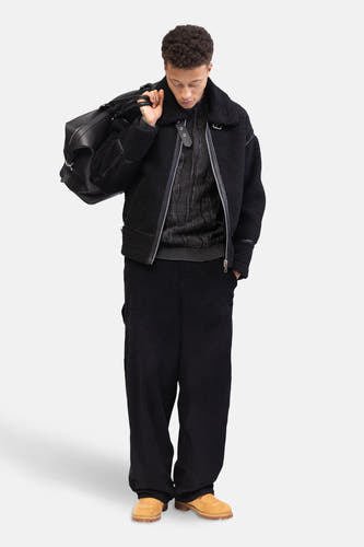 Pantalon noir de REDEFINED REBEL, en tissu corduroy, porté avec un pull, une veste et un sac à main en cuir.