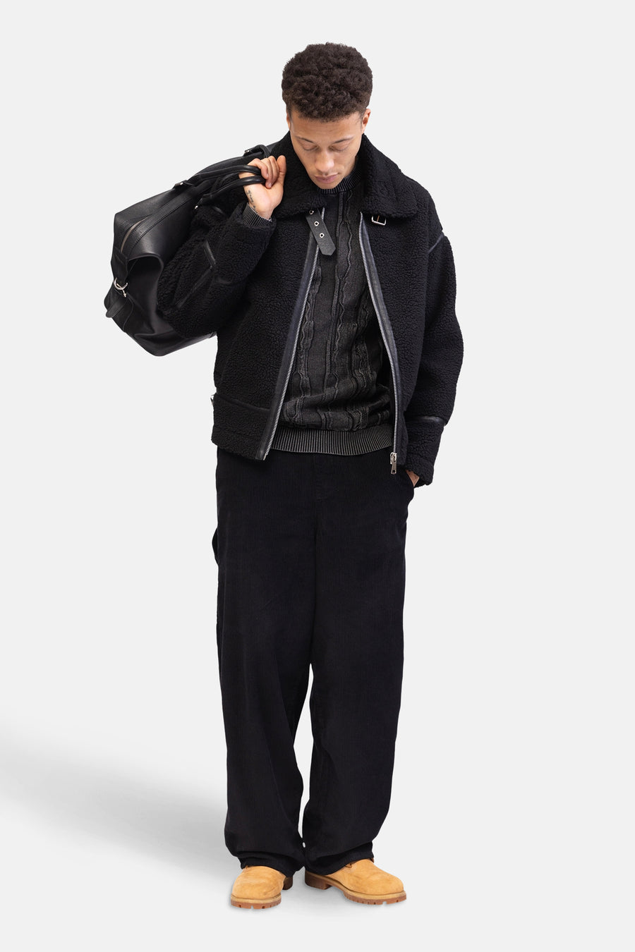 Pantalon noir de REDEFINED REBEL, en tissu corduroy, porté avec un pull, une veste et un sac à main en cuir.