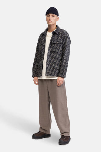 Bruine broek van REDEFINED REBEL, gecombineerd met een bedrukt jack, een wit t-shirt, een gouden ketting en zwarte schoenen.
