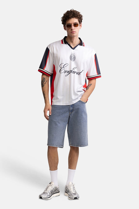 Multicolor polo met witte basis, rode en donkerblauwe panelen, van REDEFINED REBEL, met Engeland-script en logo, gedragen met denim shorts en zilveren sneakers.
