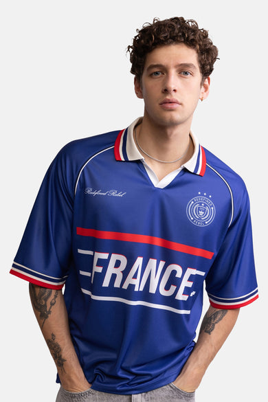 Blauwe polo met witte en rode accenten van REDEFINED REBEL, met FRANCE-print en logo, gedragen door model met tatoeages
