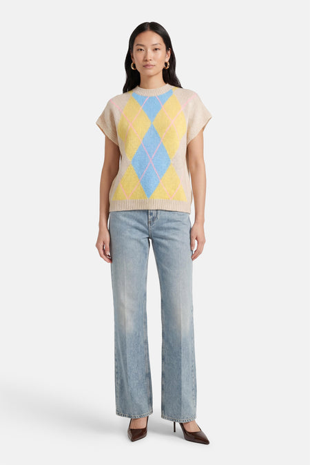 Beige mouwloos gilet van Terra di Siena, met een argyle-ruitpatroon in blauw en geel, gedragen over een jeans met wijde pijpen en bruine pumps.

