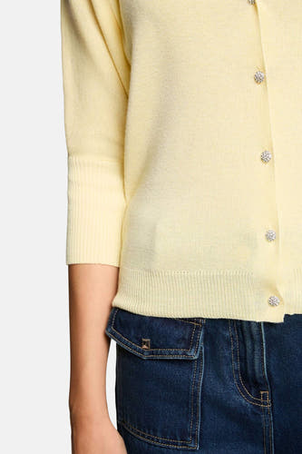 Gele fijngebreide cardigan van Morgan De Toi met driekwart mouwen, geribbelde mouw- en onderboord en glinsterende knopen, deels over een donkerblauwe denim broek.