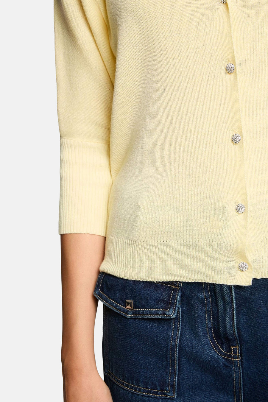 Gele fijngebreide cardigan van Morgan De Toi met driekwart mouwen, geribbelde mouw- en onderboord en glinsterende knopen, deels over een donkerblauwe denim broek.