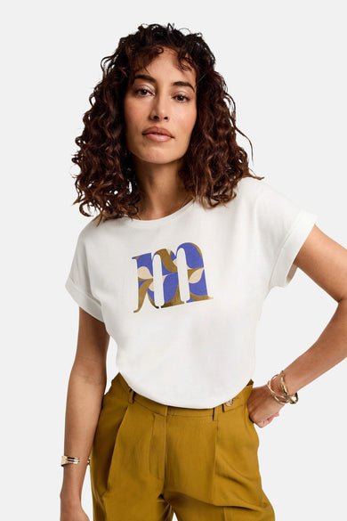 Wit T-shirt met korte mouwen van Morgan De Toi, met een grote blauwe en goudkleurige "M" print, gecombineerd met een mosterdkleurige pantalon en gouden armbanden.
