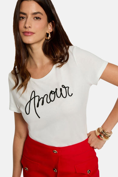 Witte top van Morgan De Toi met zwarte 'Amour' belettering, gecombineerd met een rode broek en gouden armbanden.
