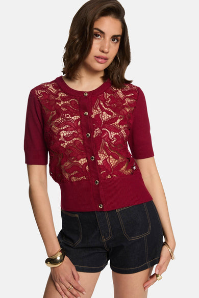 Pull rouge à col rond de Morgan De Toi, avec des bordures et des manches courtes, associé à des shorts en denim foncé et des bracelets dorés.