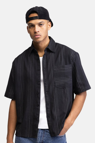 Zwart overhemd met korte mouwen van REDEFINED REBEL, met witte strepen en borstzak, gestyled met een wit T-shirt, gouden ketting, jeans en een corduroy pet.