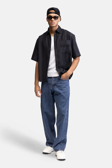 Zwart overhemd met korte mouwen van REDEFINED REBEL, met fijne witte verticale stippellijnen, open over een wit T-shirt gedragen en een blauwe spijkerbroek.