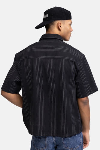 Zwart hemd met korte mouwen van REDEFINED REBEL, met dunne witte pinstripes, van achteren gezien op een model met een zwarte corduroy pet en blauwe jeans.
