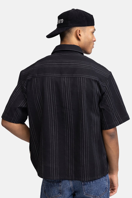 Zwart hemd met korte mouwen van REDEFINED REBEL, met dunne witte pinstripes, van achteren gezien op een model met een zwarte corduroy pet en blauwe jeans.