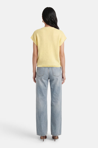 Geel gilet van Terra di Siena, met korte mouwen en een fijngebreide textuur, gecombineerd met blauwe jeans en bruine hakken, gezien van achteren.

