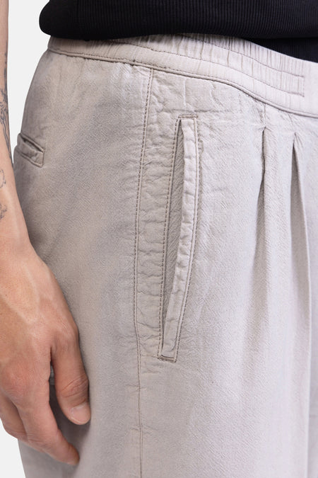 Ecru broek van REDEFINED REBEL met elastische tailleband, verticale steekzak en plooien aan de voorkant. Links een deel van een arm met tatoeages.