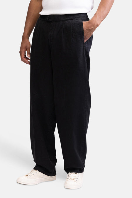 Pantalon noir de REDEFINED REBEL avec coupe ample, taille élastique et poche latérale, porté avec un t-shirt blanc et des baskets beiges.