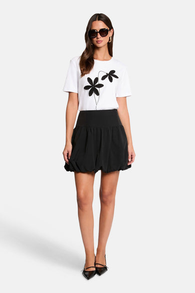 Witte T-shirt met korte mouwen van Morgan de Toi, met zwarte bloemenprint, gecombineerd met een zwarte ballonrok en zwarte schoenen.