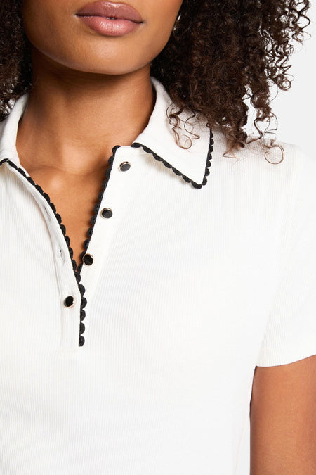 Close-up van een witte ribgebreide polo met zwarte bies en knopen, gedragen door een persoon met donkere krullen.