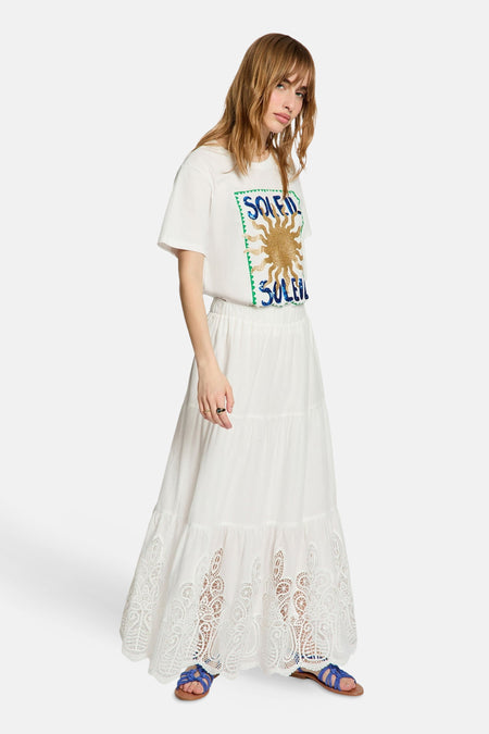 T-shirt blanc Morgan De Toi avec imprimé 'SOLEIL', soleil à paillettes dorées et lettres à sequins bleus, porté avec une longue jupe blanche et des sandales bleues.