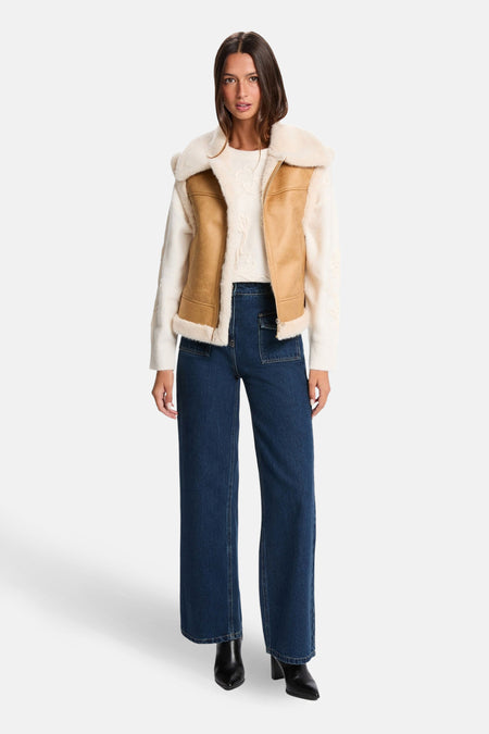 Vrouw in beige vest, crèmekleurige trui, blauwe jeans en zwarte laarzen, staand.