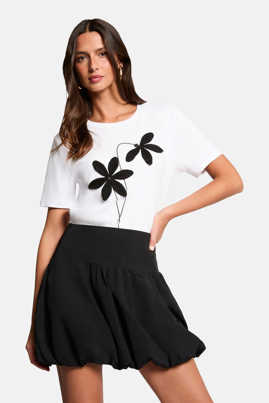 Jupe courte noire de Morgan De Toi, avec une coupe ballon et associée à un t-shirt blanc avec des applications de fleurs noires.