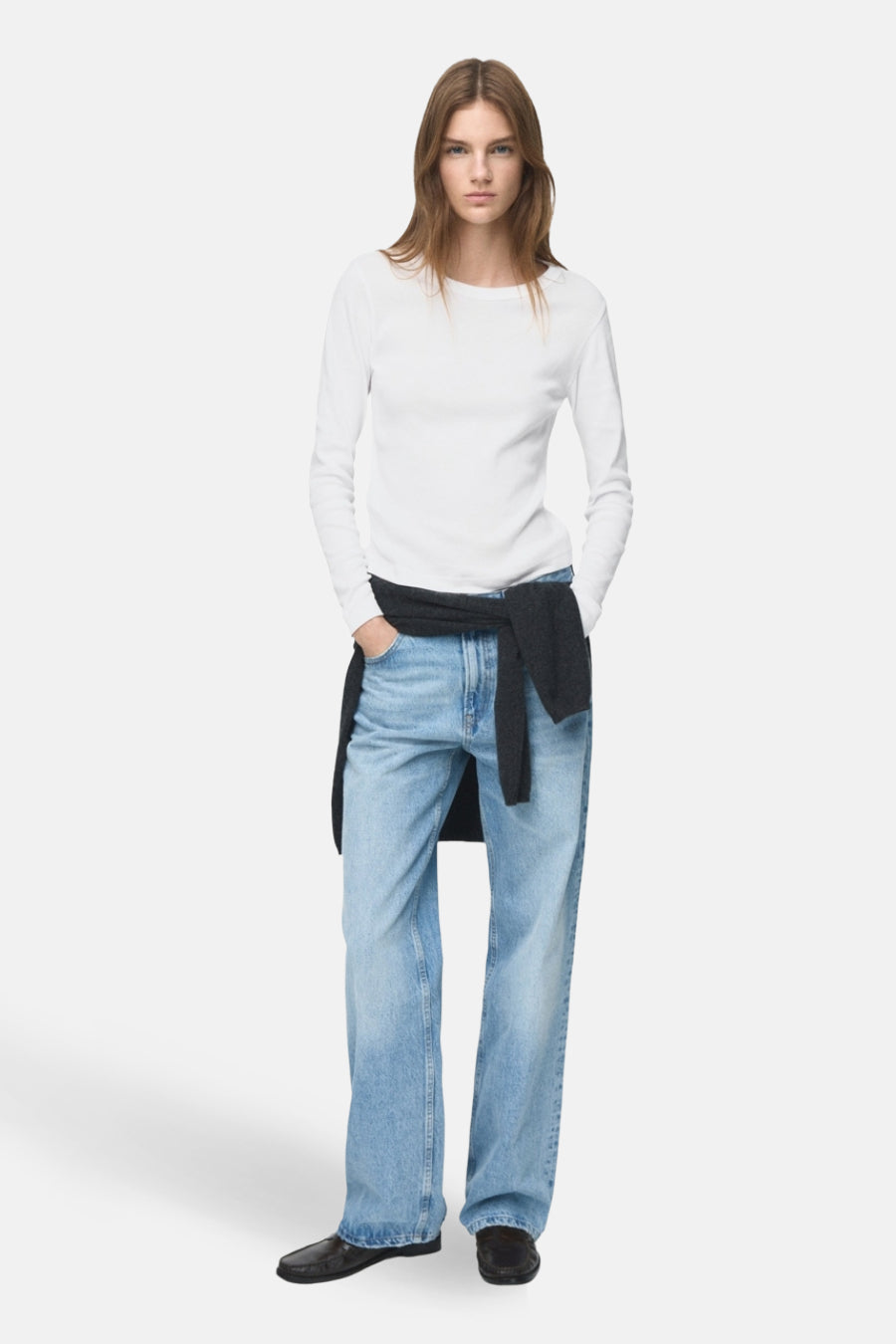 T-shirt blanc à manches longues de MANGO, porté avec un pull sombre autour de la taille et des jeans larges.