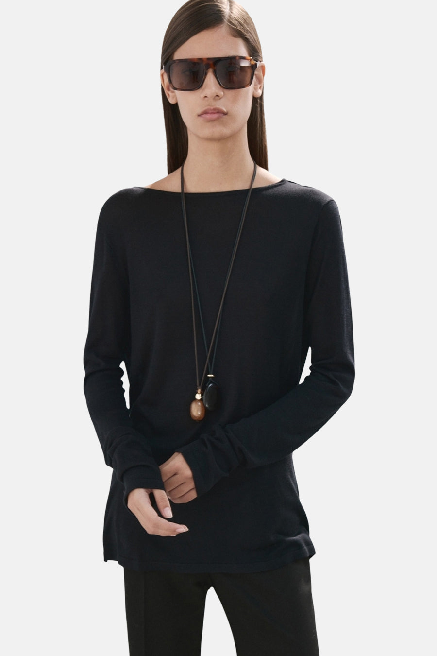 Pull noir à col rond de MANGO, à manches longues et à encolure simple, porté avec un long collier et des lunettes de soleil.