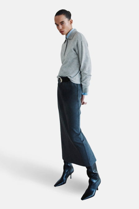 Vrouw in grijze trui, lange rok met riem en zwarte laarzen.