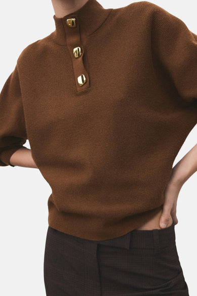 Bruine trui met rolkraag van MANGO, afgewerkt met gouden knopen aan de voorkant en gecombineerd met een geruite broek.
