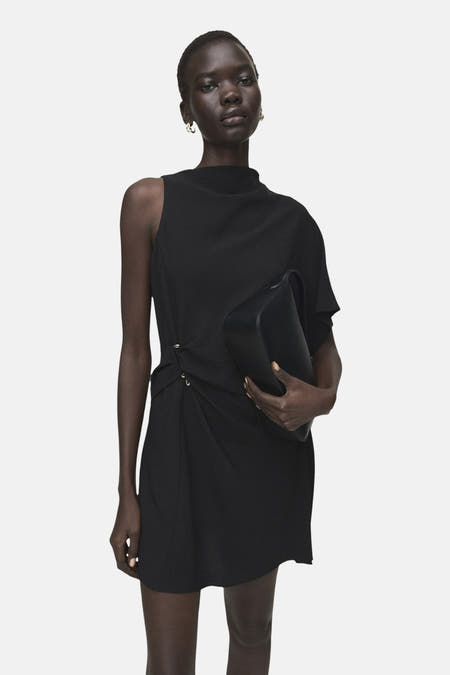 Zwarte mouwloze jurk van MANGO, met een asymmetrisch ontwerp en draperingen aan de zijkant, gecombineerd met een zwarte tas.