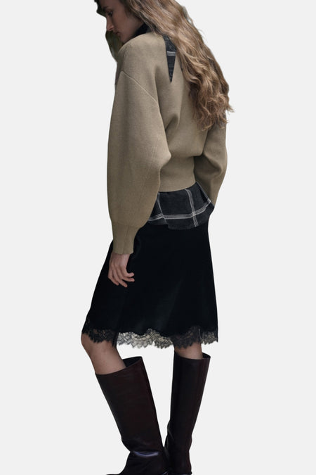 Vrouw in kaki trui, geruite blouse, zwarte rok met kant, en bruine laarzen.