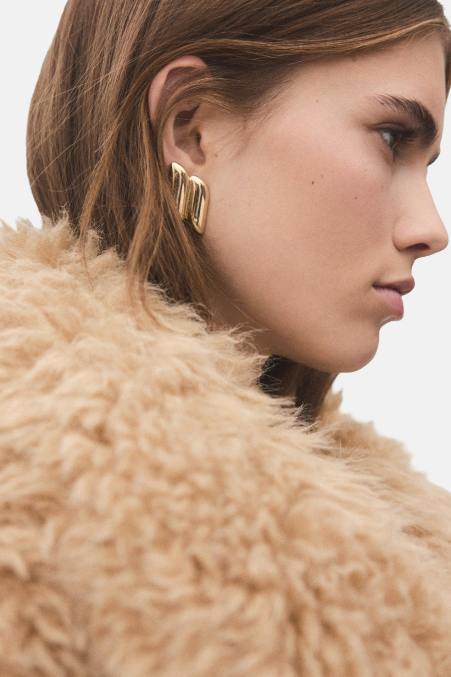 Boucles d'oreilles en or de MANGO, portées avec une veste douce et beige dans un profil latéral.